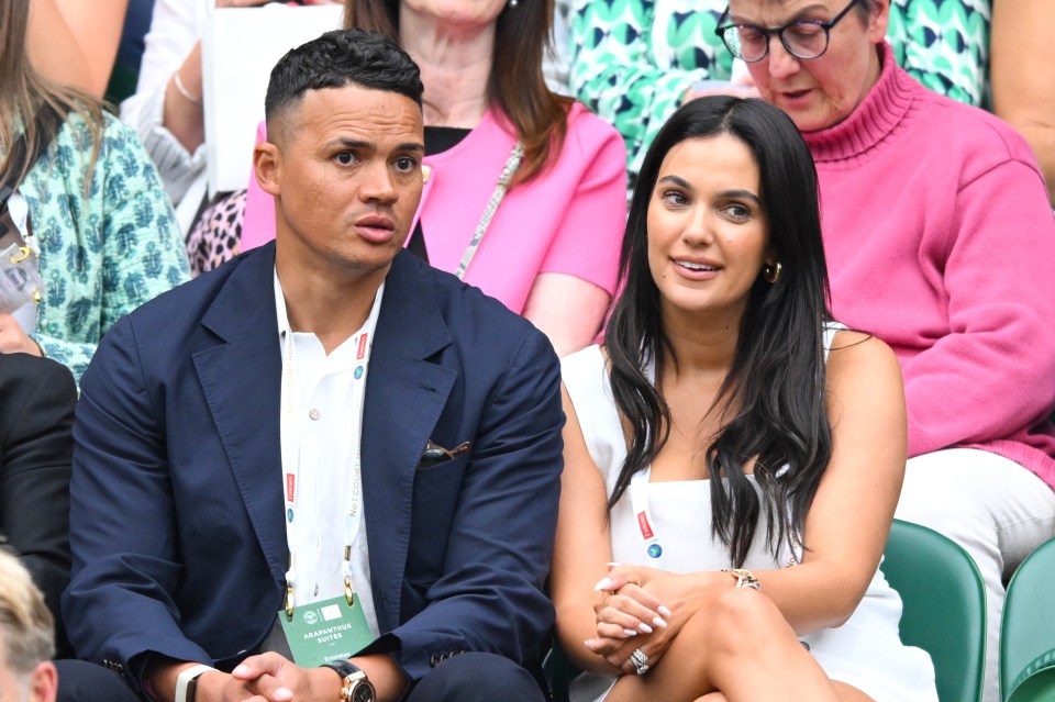 a man and a woman sit in a stadium with the man wearing a wimbledon badge