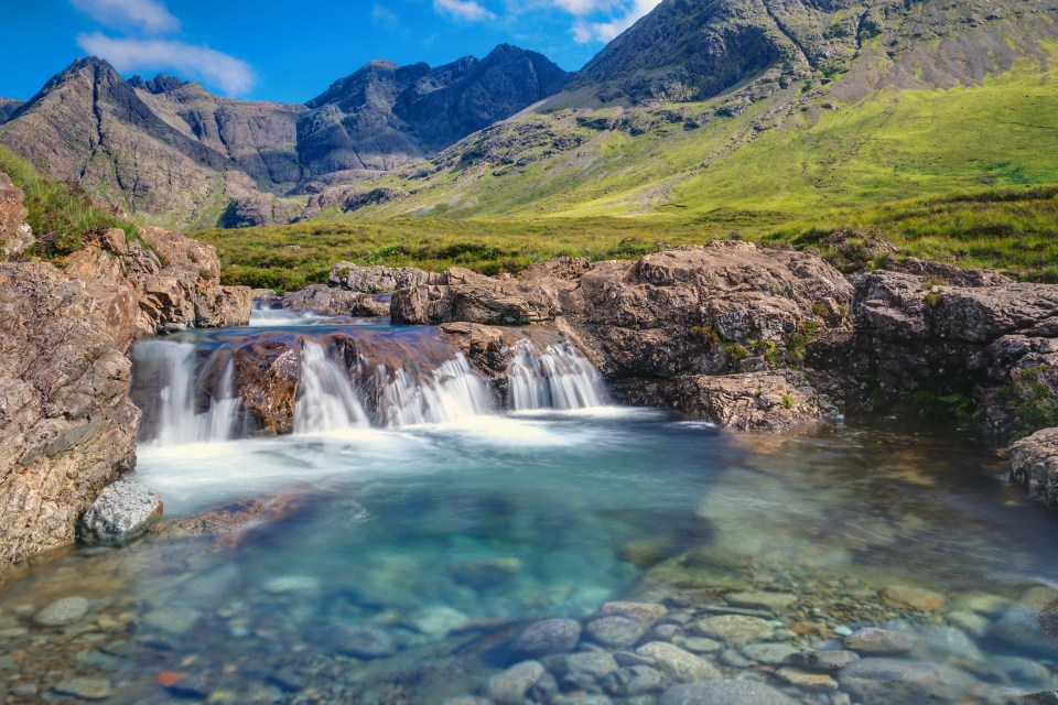 a waterfall surrounded by rocks and grass with mountains in the background