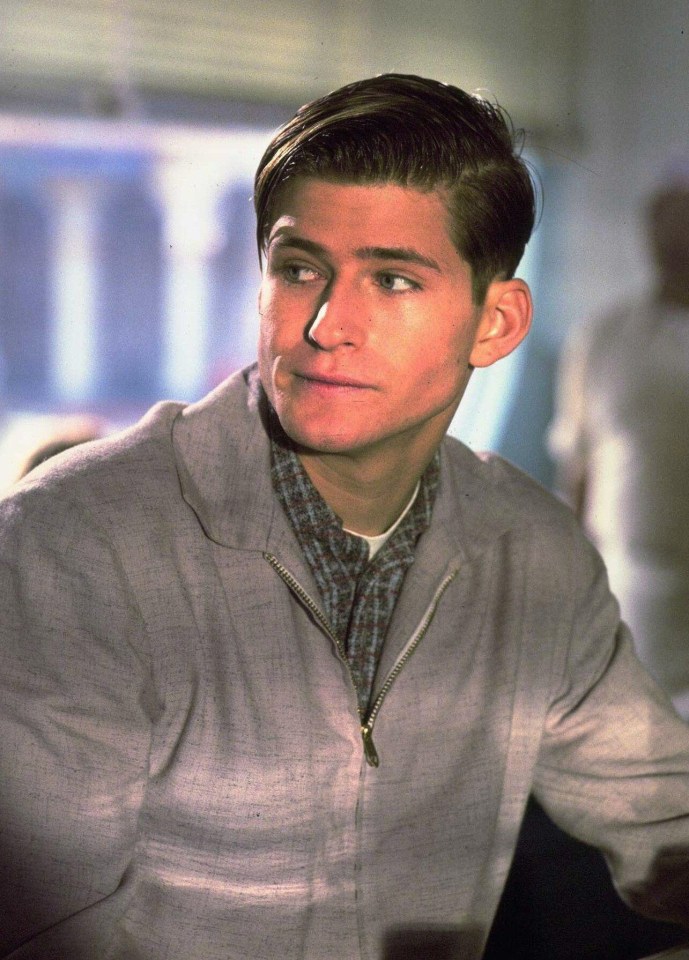 a man in a grey jacket and plaid shirt looks at the camera
