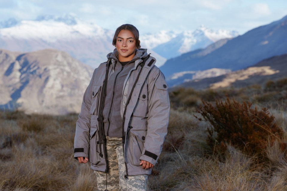 a woman in a grey jacket stands in a field with mountains in the background