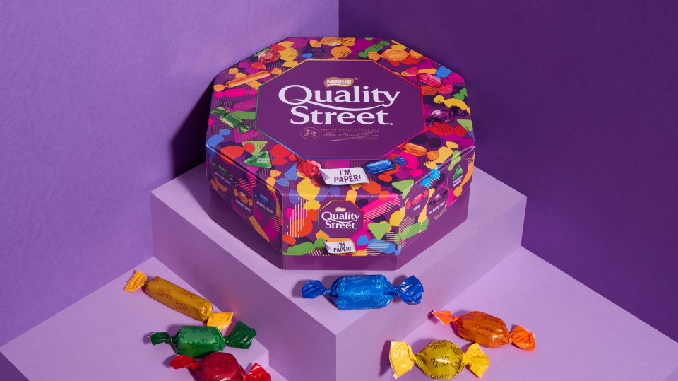 a box of quality street candy sits on a purple surface