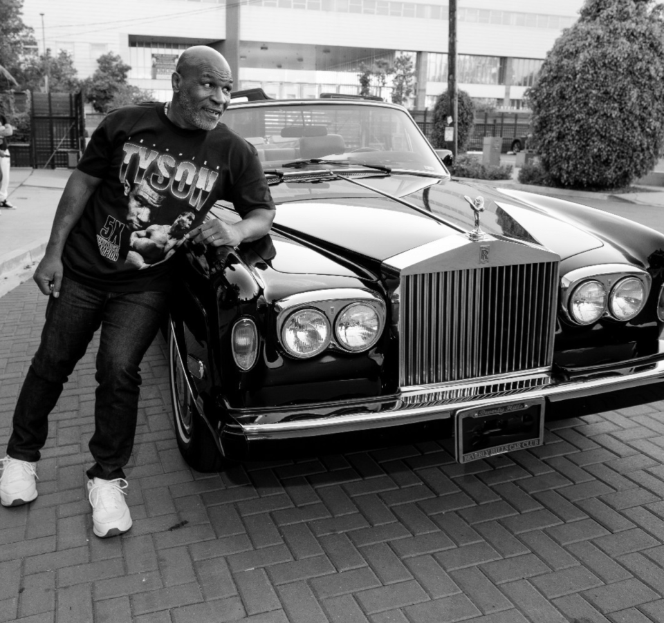a man wearing a tyson shirt leans against a black car