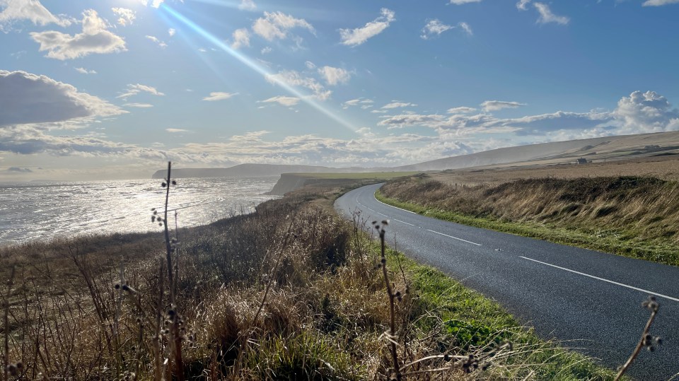 Photo taken in august 2023 in Brook, Isle of Wight, UK.its late afternoon and the sun is setting, to the left of the photo the sea is visible, followed by the grassy meadow and the asphalt road winds around the corner. Cliffs visible. Reflections in the water. Dry grasses in the foreground