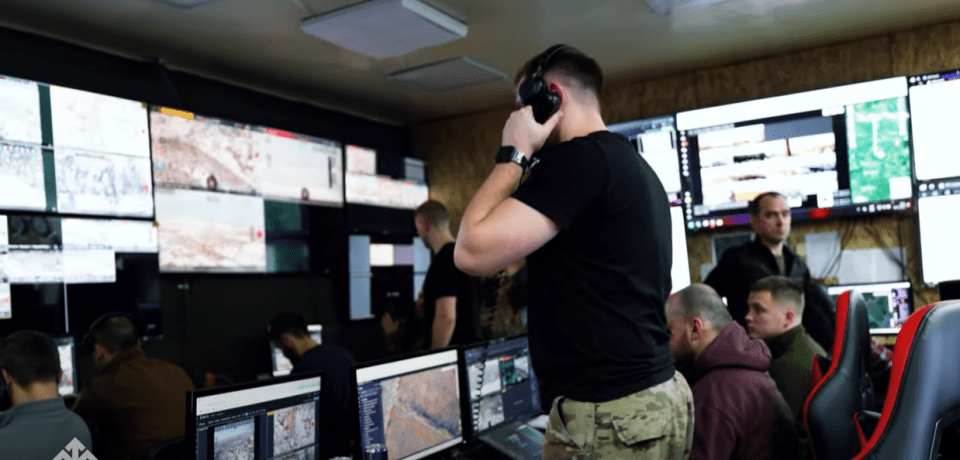 A control room with multiple screens displaying battlefield information; a person in headphones adjusts their headset.