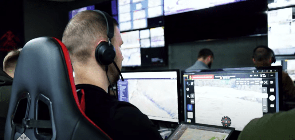 A person wearing a headset monitors computer screens displaying a robot battle.