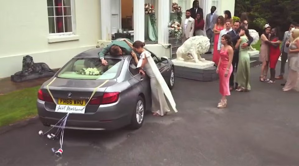 A newlywed couple by their car, surrounded by wedding guests.