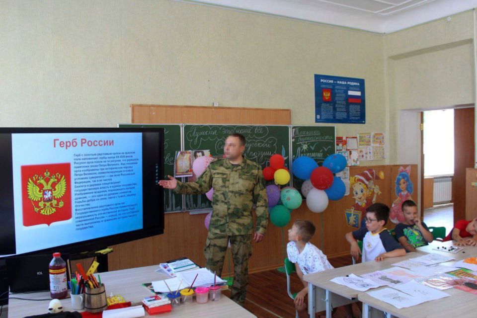 A man in uniform gives a presentation about the Russian coat of arms to a group of children.