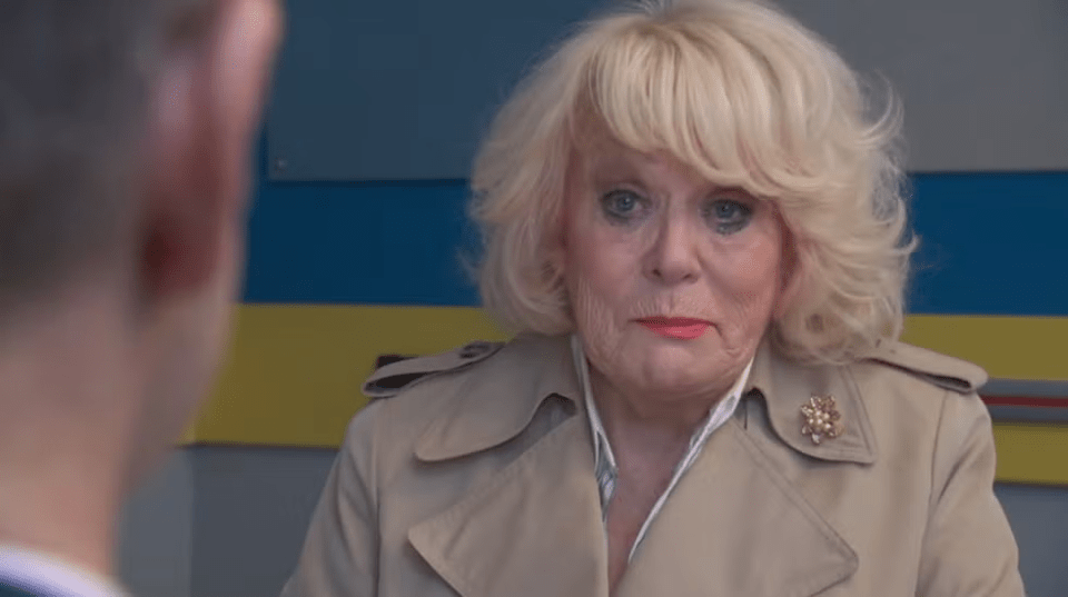 Older woman in beige coat speaking to someone.