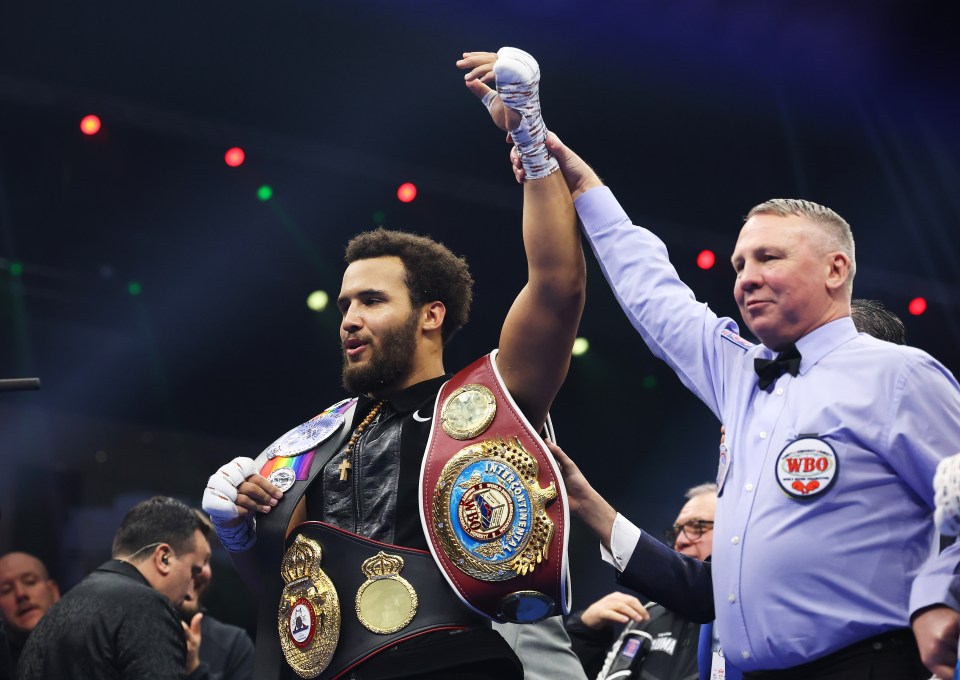 Moses Itauma raises his hand in victory after a boxing match, wearing multiple championship belts.
