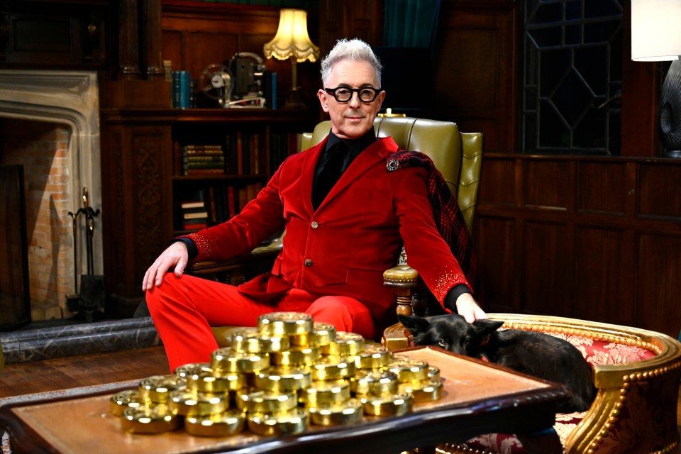 Alan Cumming in a red suit sits in a chair with a dog, a pile of gold tins on a table in front of him.