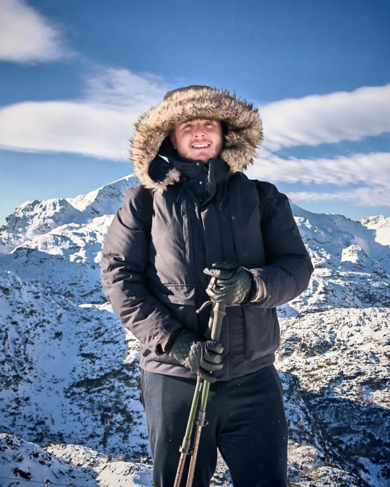 Smiling man in winter coat and gloves holding trekking poles in snowy mountains.
