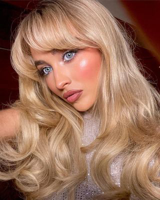 Woman with long blonde wavy hair and bangs.