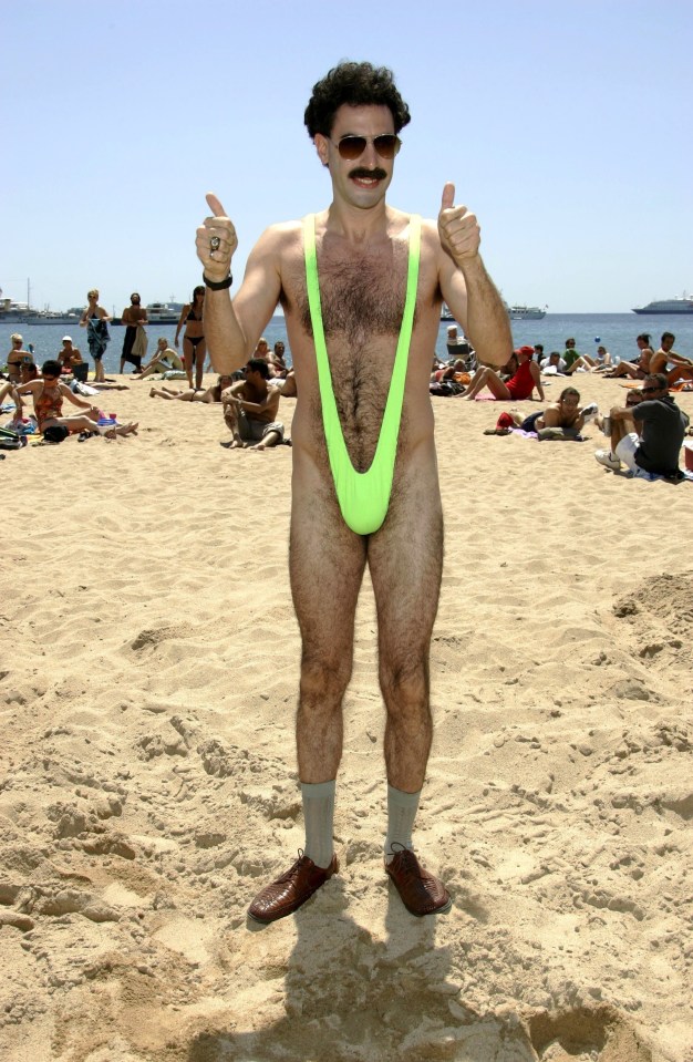 Sacha Baron Cohen as Borat giving a thumbs-up on a beach.