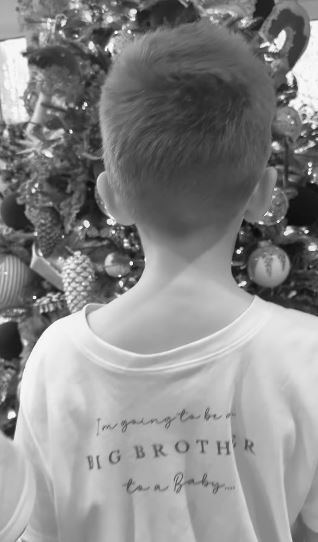 Boy in white shirt that says "I'm going to be a Big Brother to a Baby..." standing in front of a Christmas tree.