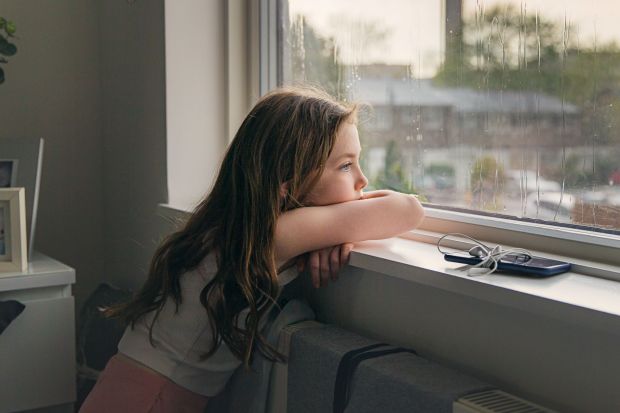 Girl looking out window, phone and earbuds on sill.