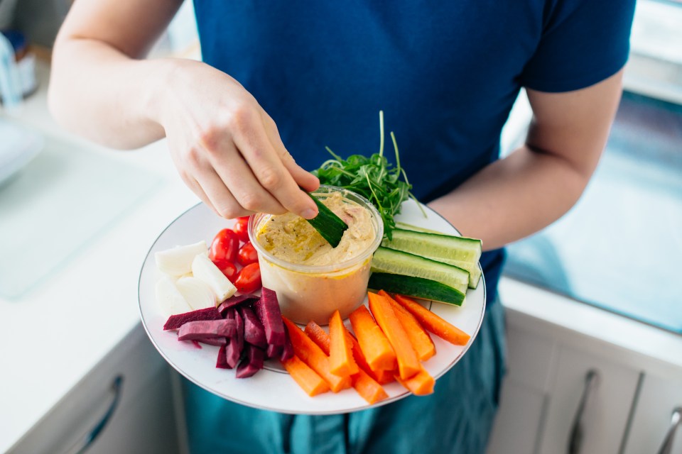 Person dipping cucumber into hummus with raw vegetables.