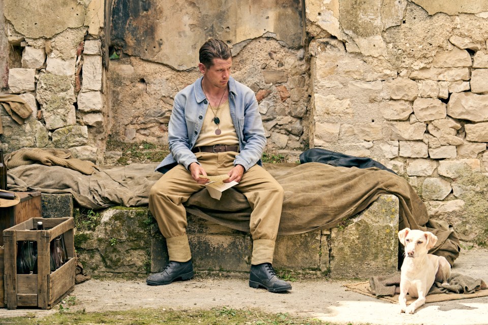 A soldier sits on a makeshift bed reading a letter, with a small dog lying nearby.