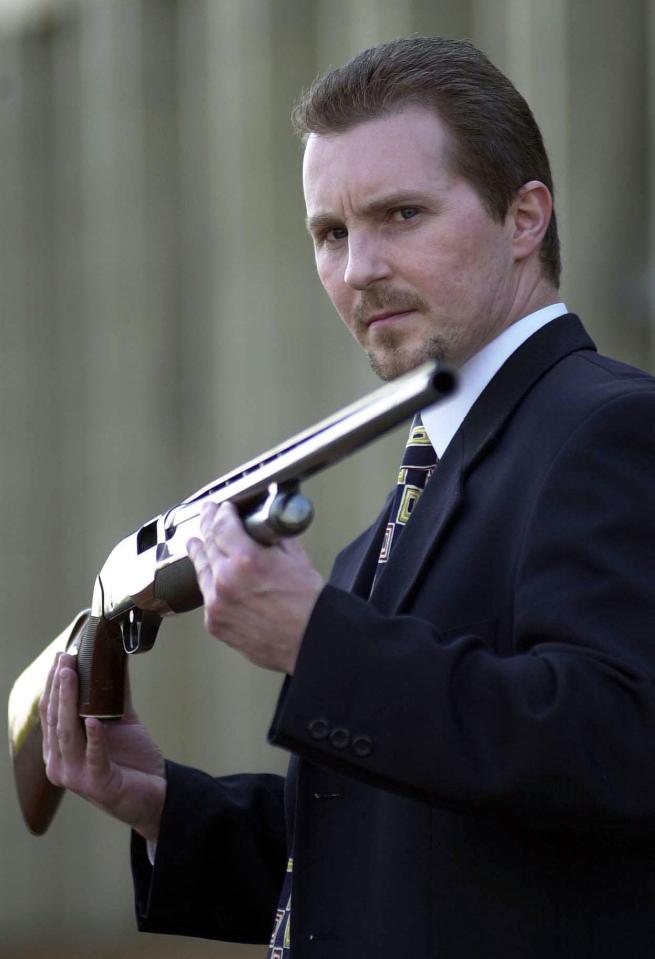 A man in a suit holds a shotgun.