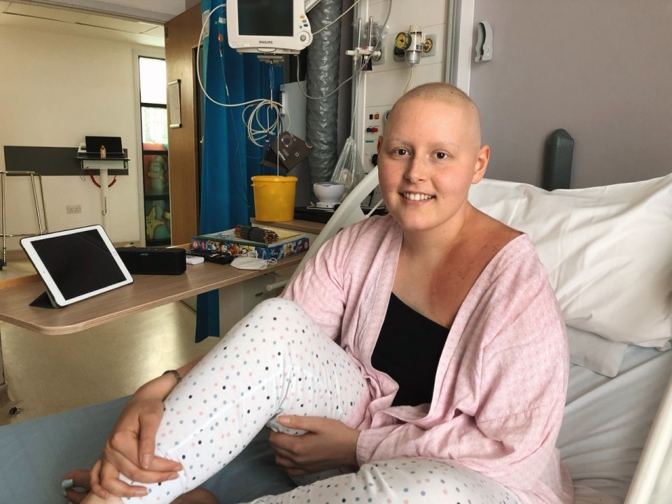 Woman with shaved head sitting in hospital bed, smiling.