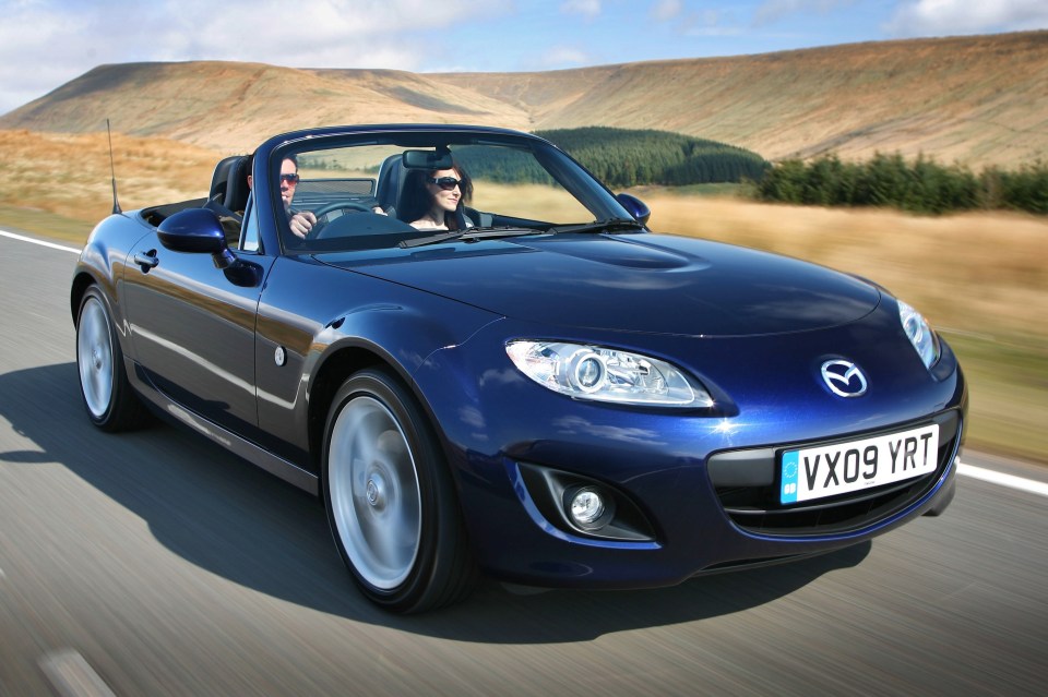 A blue Mazda MX-5 convertible driving on a road.