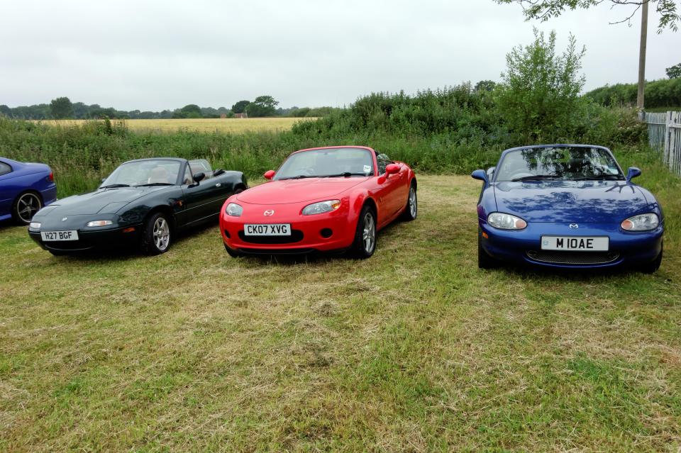 Three generations of Mazda MX-5 Miata parked in a field.
