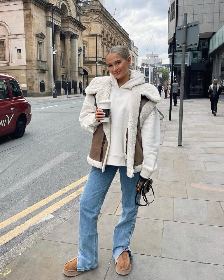 Woman in Ugg boots, jeans, and a sherpa vest holding a coffee cup.