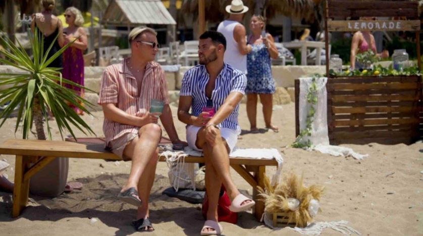 Two men sitting on a bench on a beach, talking.  A lemonade stand is visible in the background.
