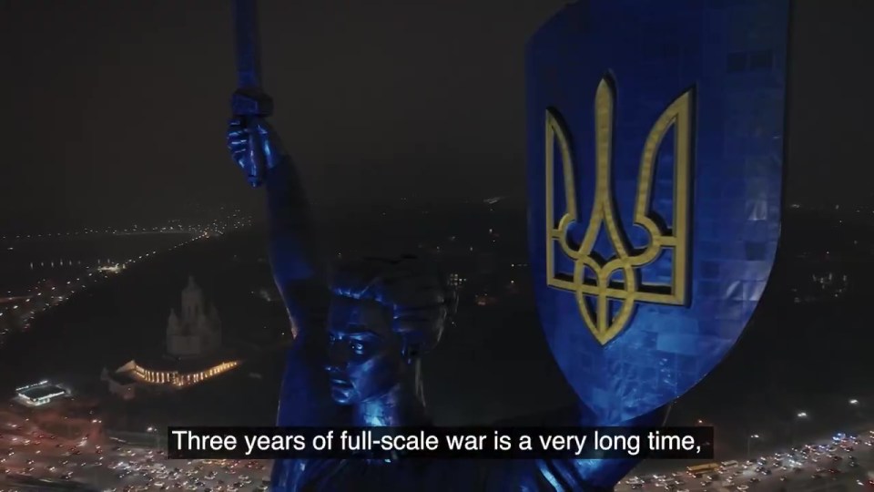 Nighttime aerial view of a statue holding a shield with the Ukrainian trident, commemorating three years of war.