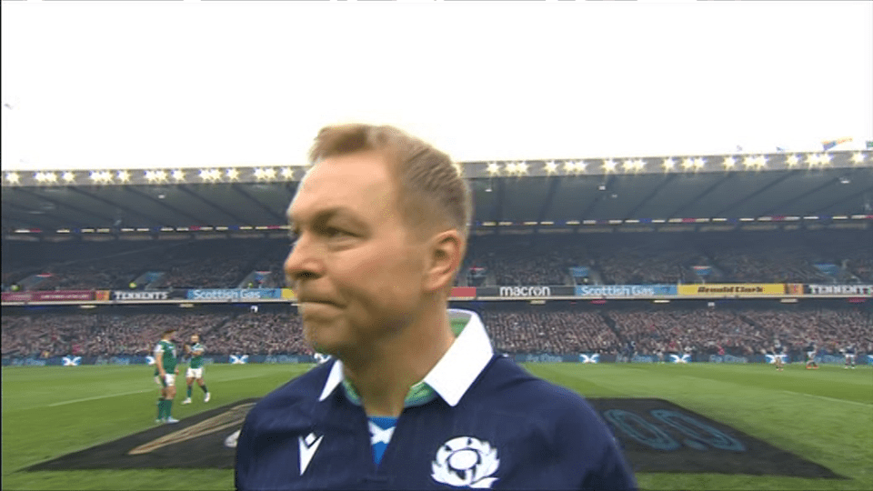 A man in a Scotland rugby jersey stands on a rugby pitch.
