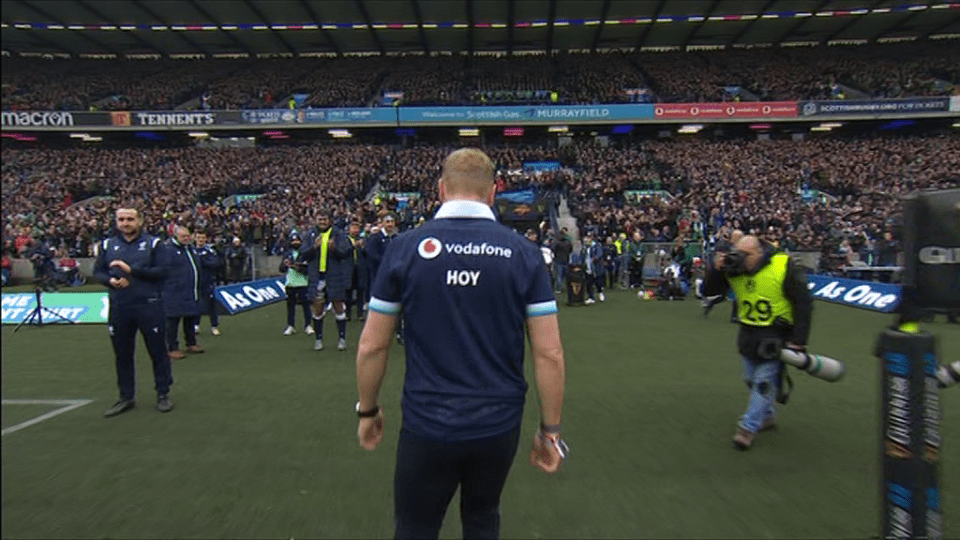 A rugby player walks across a field after a game, with a large crowd cheering in the background.