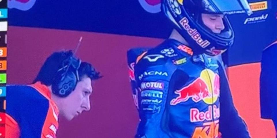 A racer in a Red Bull racing suit and helmet stands next to a person wearing headphones.