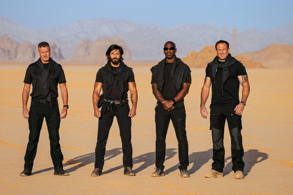 Four men in black tactical gear stand in a desert.