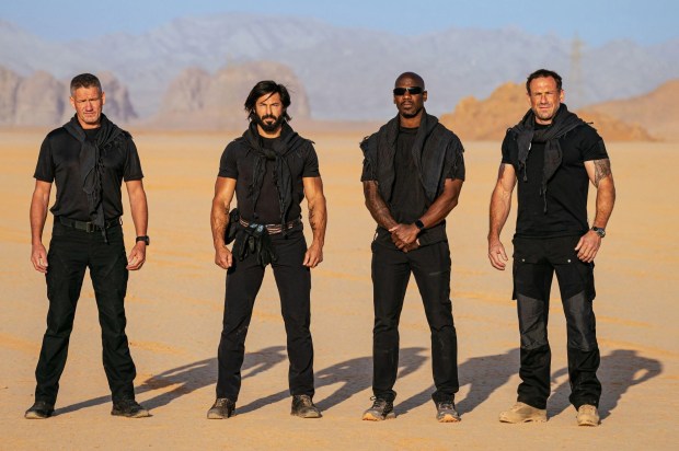 Four men in black tactical gear stand in a desert.