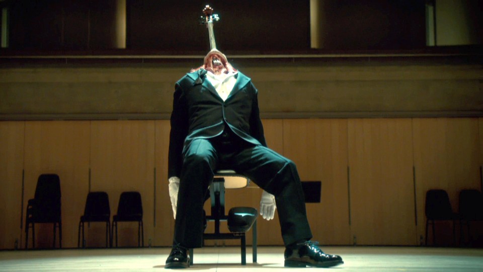 A headless man in a tuxedo sits on a chair with a cello protruding from his neck.