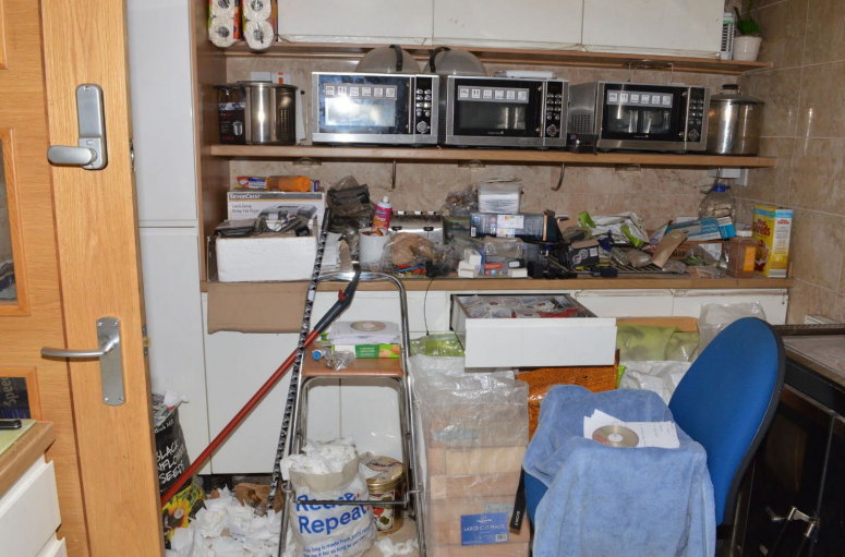 A cluttered office with microwaves, shelves, and various items on the floor.