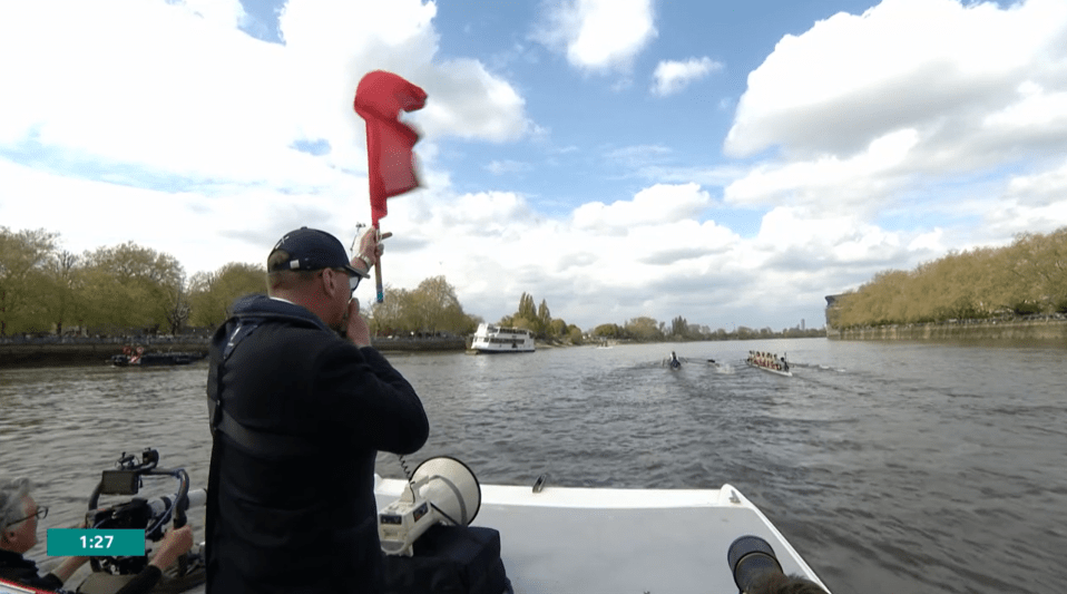 A boat race official signals from a boat, with rowers in the background.