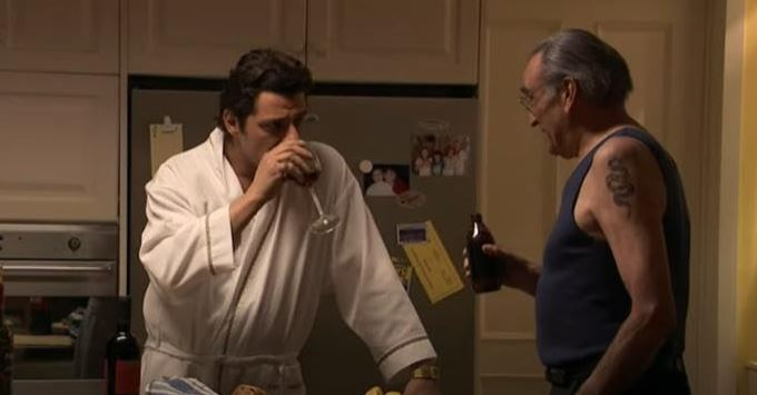 Two men in a kitchen; one in a bathrobe drinks wine, the other holds a beer bottle.