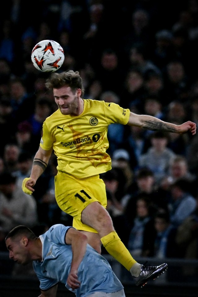 Soccer player in yellow jersey jumping for the ball, another player in light blue jersey below.