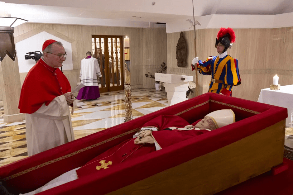A cardinal standing over an open casket containing a deceased pope.  A Swiss Guard stands nearby.
