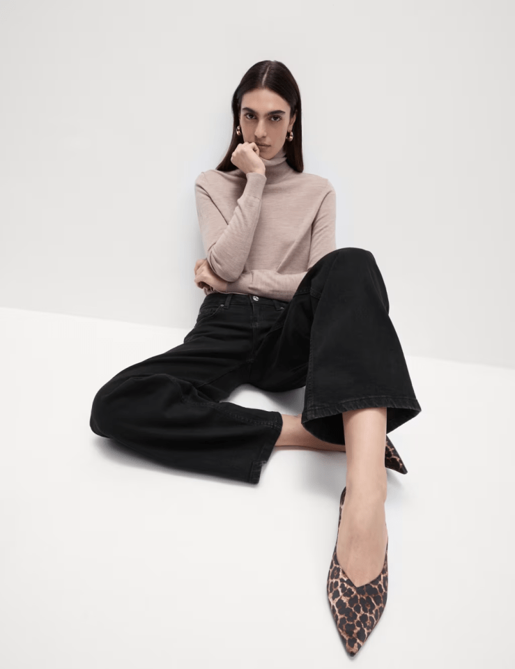 Woman sitting on the floor wearing a beige turtleneck, black jeans, and leopard-print shoes.