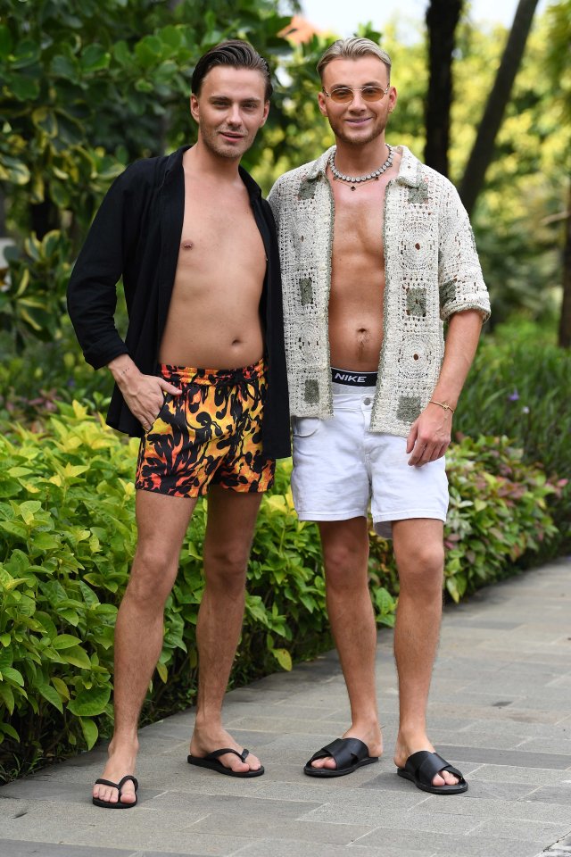 Two shirtless men in swim trunks and sandals stand together outdoors.