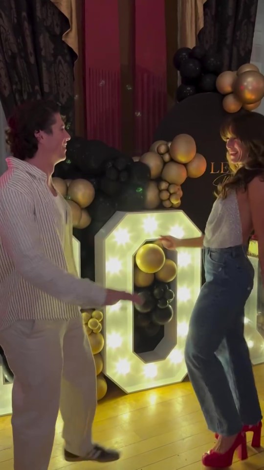 Two people dancing in front of a lighted "30" made of balloons.
