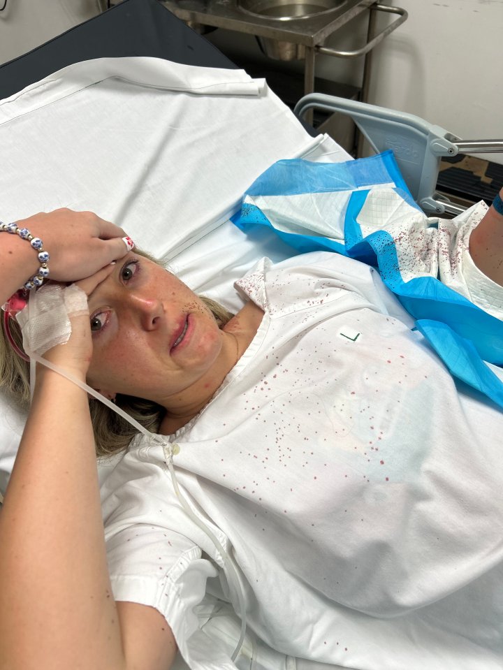 Woman in hospital gown with blood stains, holding her head.