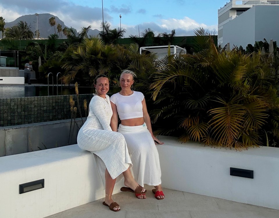Two sisters in white dresses sitting on a wall by a pool.