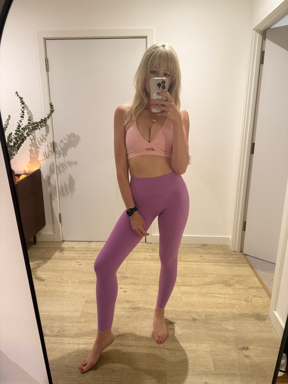 Woman in purple leggings and pink sports bra taking a selfie in a mirror.