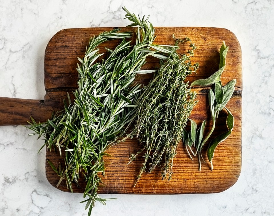 Rosemary, thyme, and sage sprigs on a wooden cutting board.