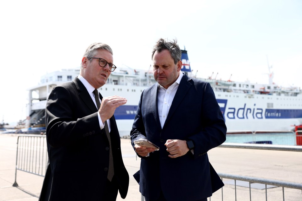 Two men in suits talking, one looking at a phone, with a ferry in the background.