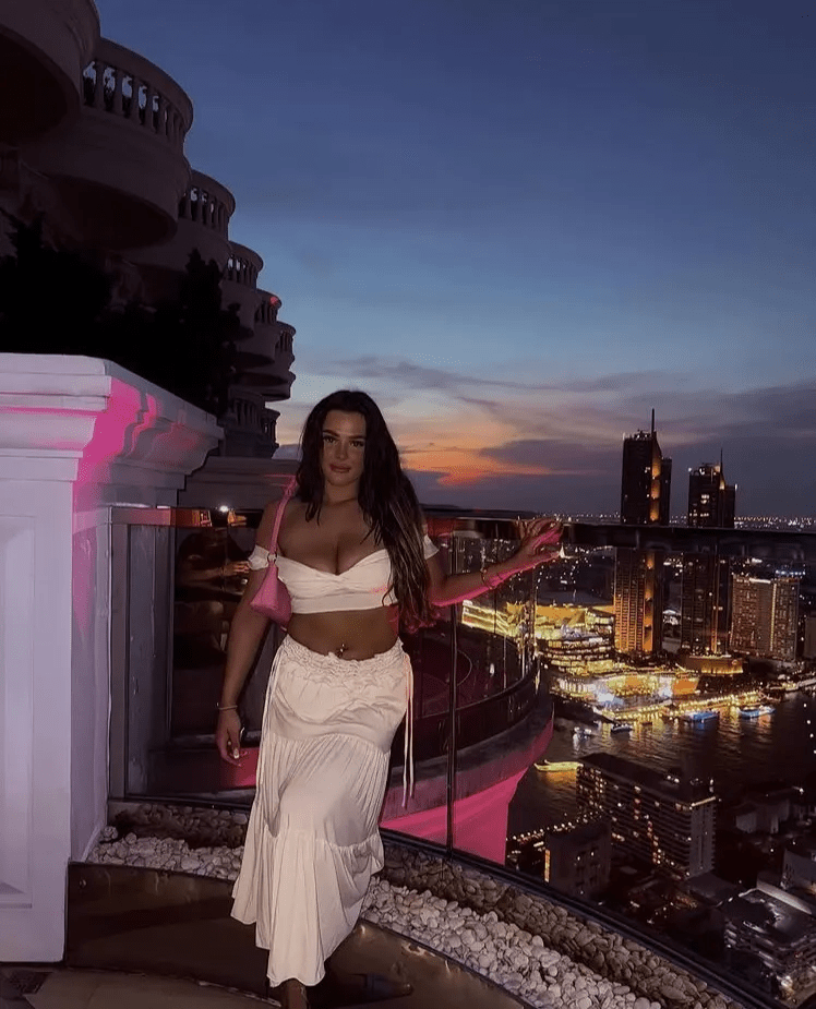 Woman in a cream-colored outfit on a rooftop overlooking a city at sunset.
