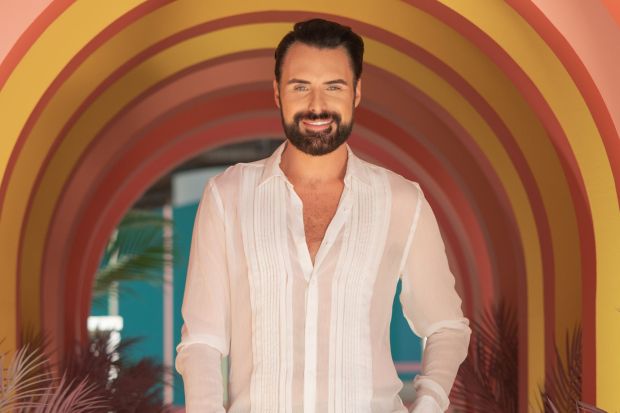 An image collage containing 1 images, Image 1 shows Rylan Clark in a white shirt and beige pants, smiling in a colorful archway