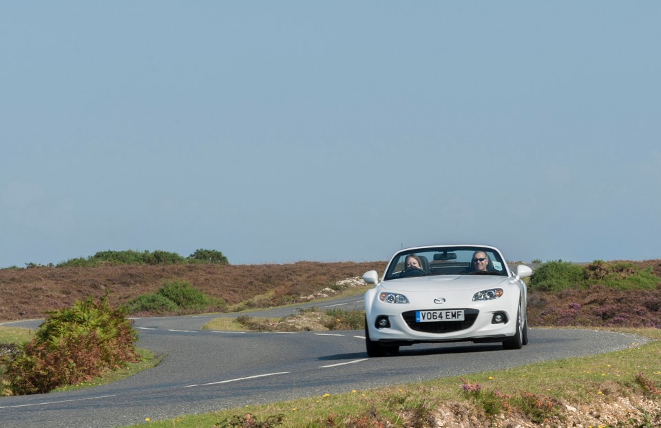 White Mazda MX-5 Roadster driving on a country road.
